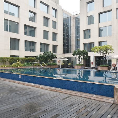 新德里国际机场假日旅馆酒店 外观 照片 The photo shows a modern outdoor swimming pool situated in a courtyard surrounded by several buildings. The pool has a sleek design with a blue tiled edge and is bordered by wooden decking. There are a few trees and plants around the area, adding som