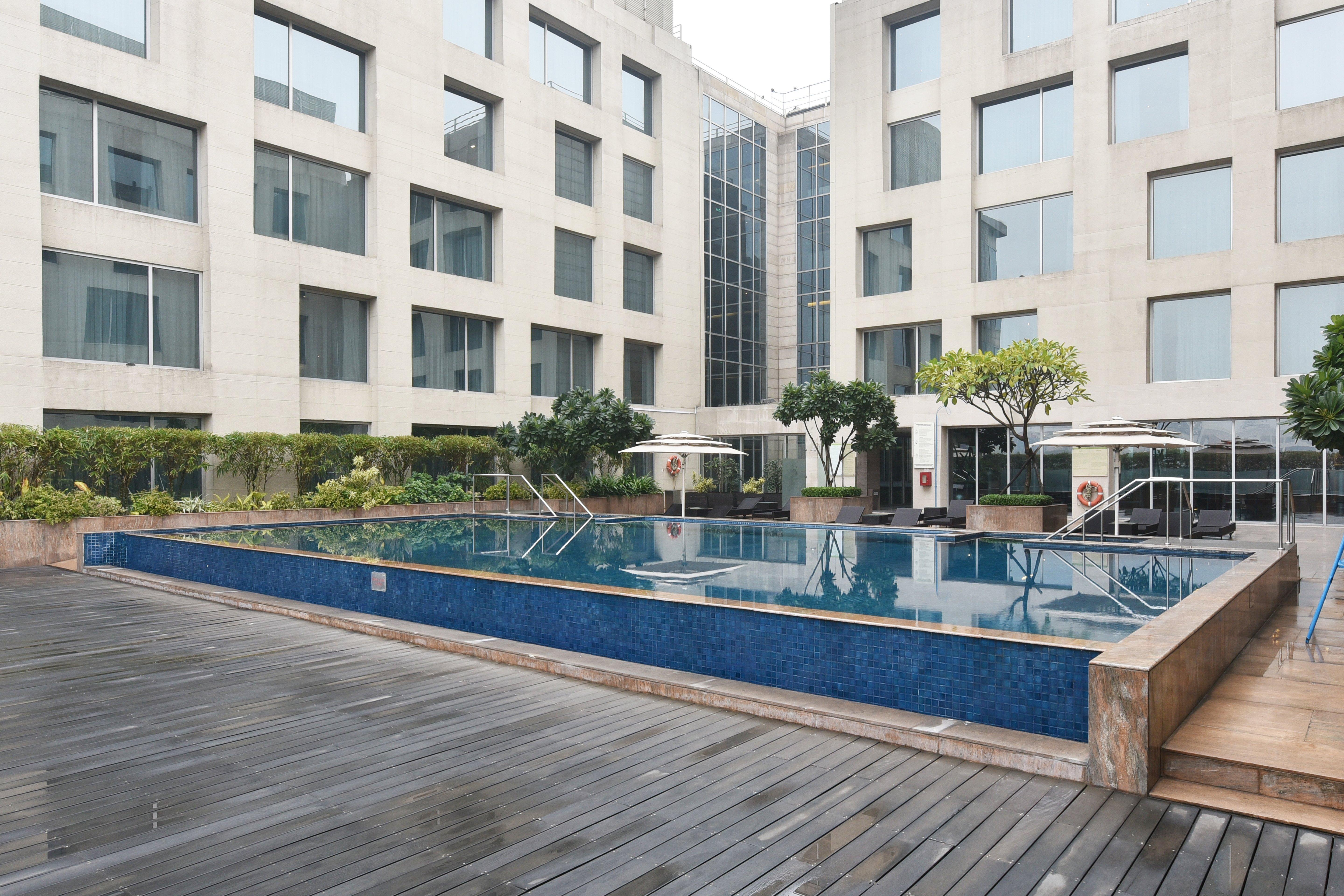新德里国际机场假日旅馆酒店 外观 照片 The photo shows a modern outdoor swimming pool situated in a courtyard surrounded by several buildings. The pool has a sleek design with a blue tiled edge and is bordered by wooden decking. There are a few trees and plants around the area, adding som