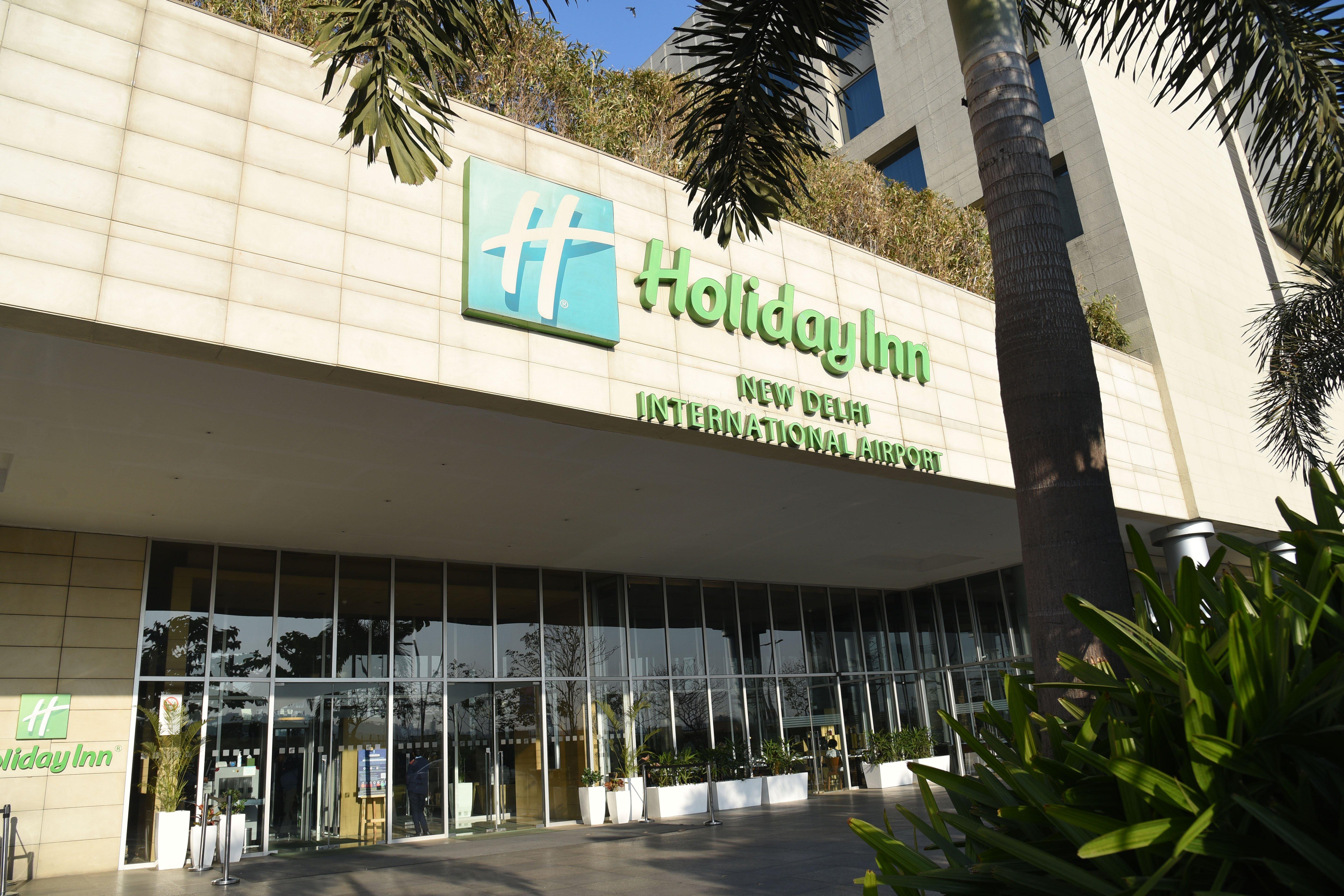 新德里国际机场假日旅馆酒店 外观 照片 The photo shows the exterior of a Holiday Inn hotel, specifically the Holiday Inn New Delhi International Airport. The building features a modern design with a prominent green and white sign displaying the hotel name. There are large glass doors at t