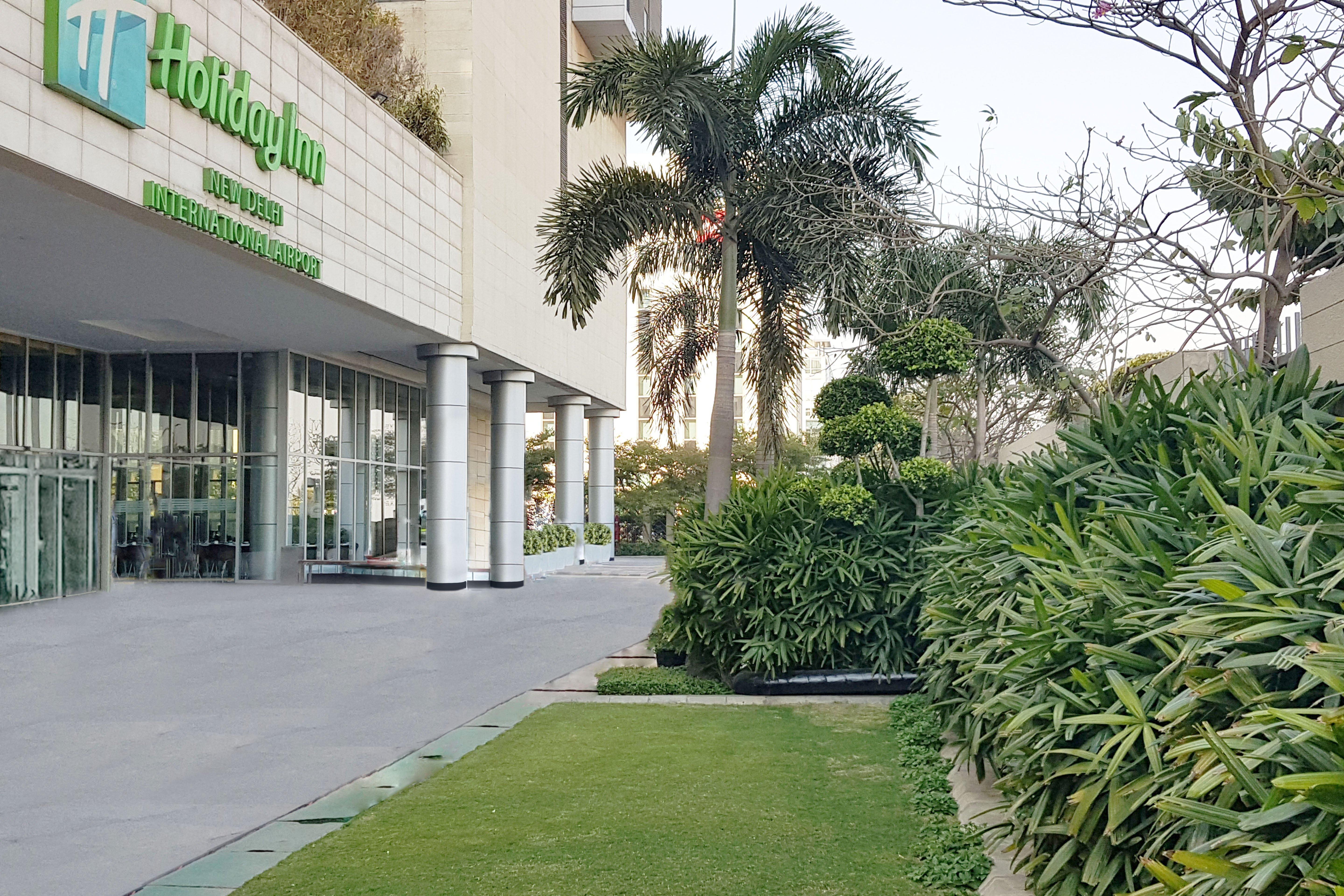 新德里国际机场假日旅馆酒店 外观 照片 The photo shows the exterior of a Holiday Inn hotel. In the foreground, there is a well-maintained green lawn and various ornamental plants, creating a pleasant garden area. The building features large glass doors and columns, and the hotel's name an